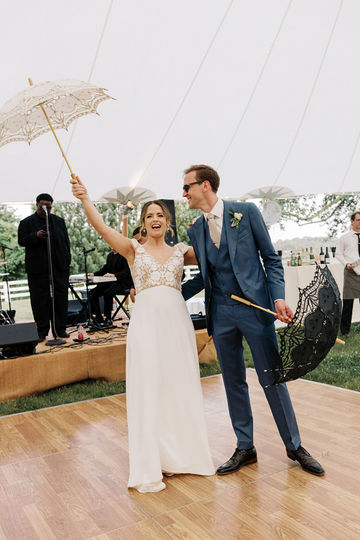 Adele and James standing in the reception tent holding parasols after their 'Second line'. Adele cheers. The band is playing on a stage behind them.
